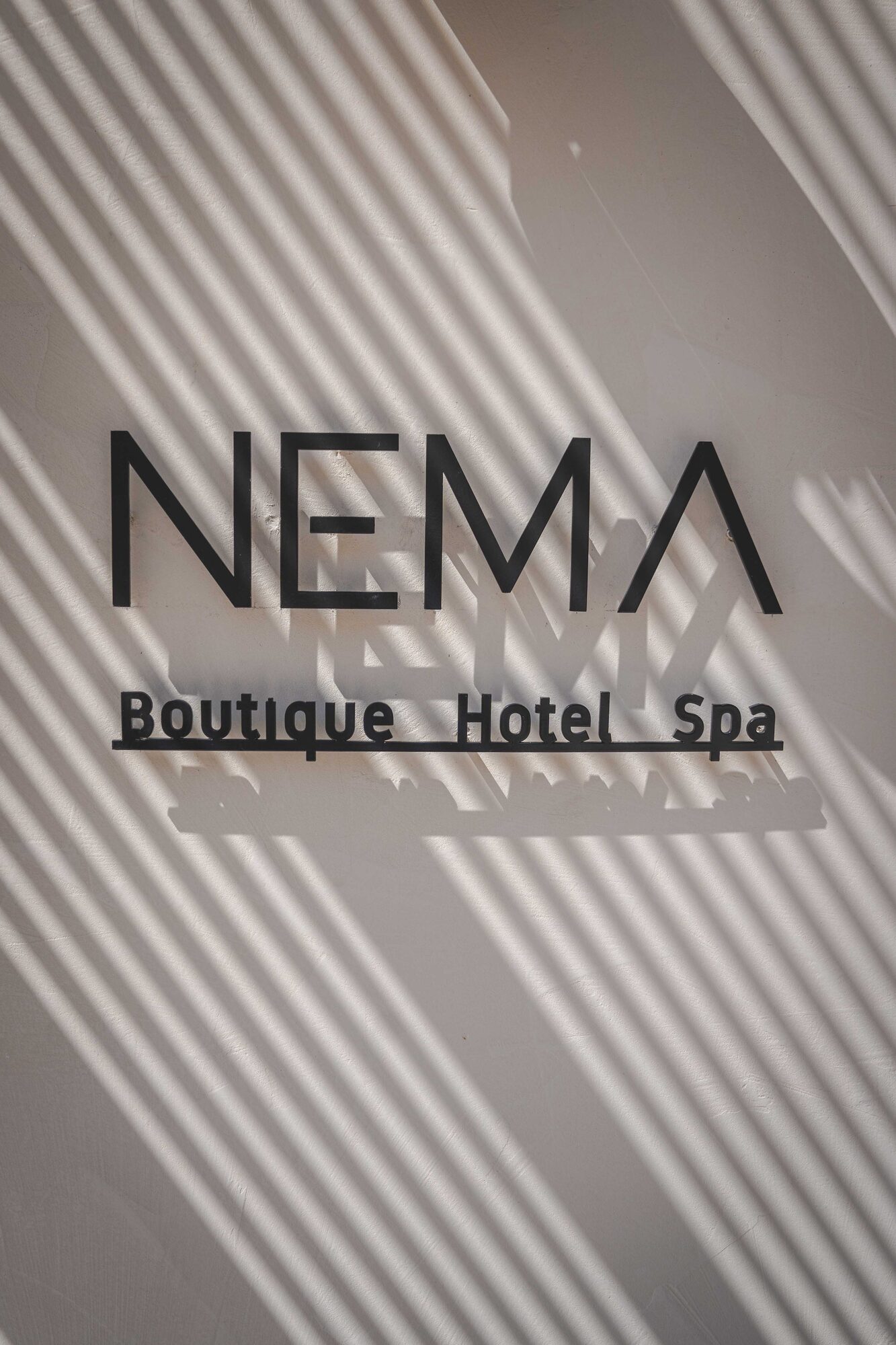 The black NEMA wordmark fixed to a pale plaster wall, crossed by even stripes of light and shadow thrown by a blind