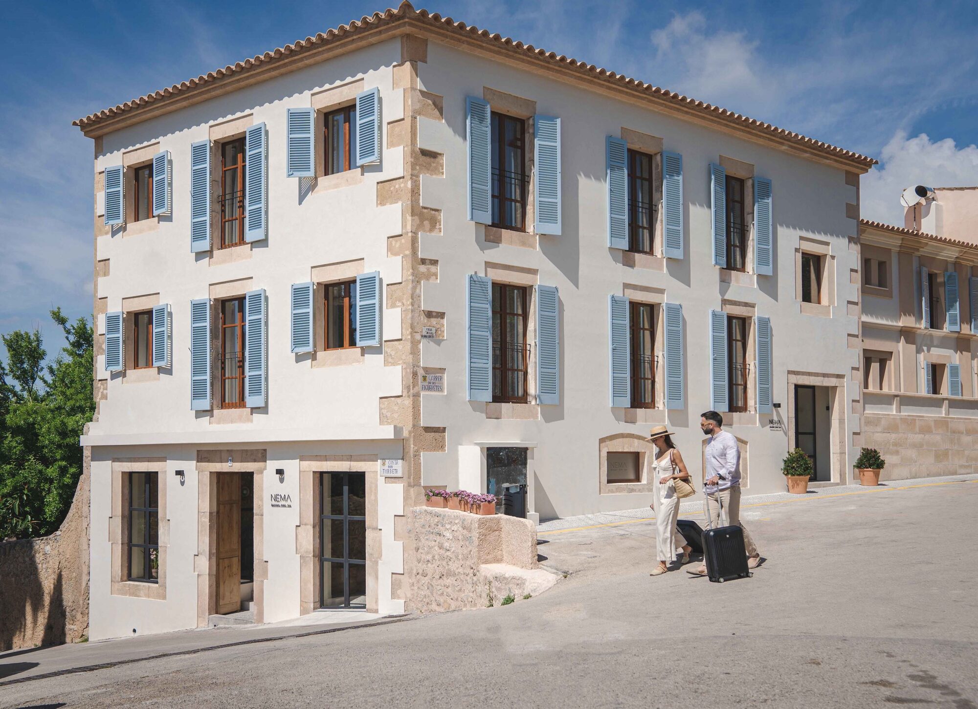 The restored 19th-century facade of NEMA on a quiet sloping street in the old town of Artà, pale rendered walls with marès-stone corners and an even row of powder-blue louvered shutters under a clear sky