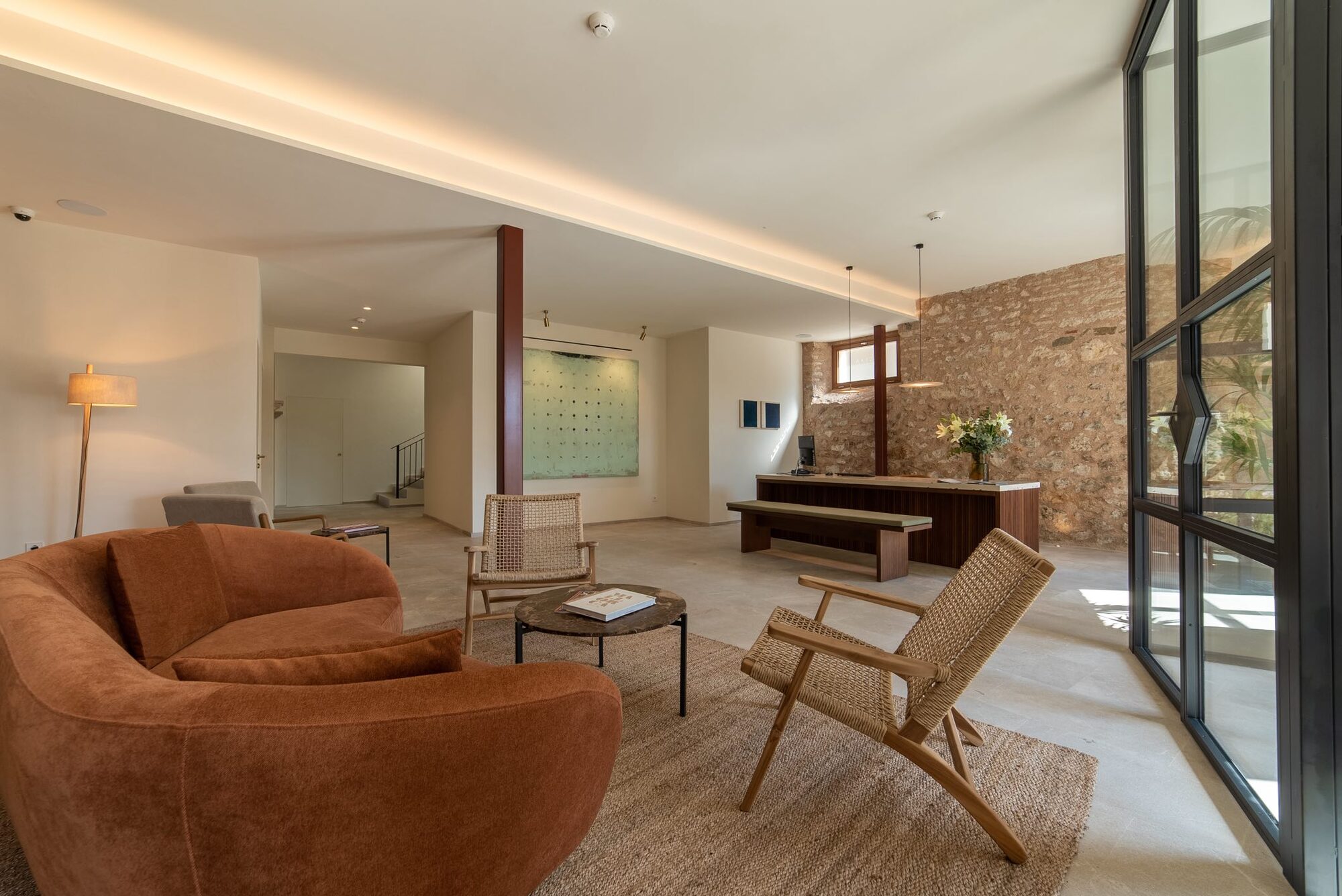 The NEMA lounge: a rust-red curved sofa and woven chairs on a jute rug, a long oak table, and an original marès-stone wall beside a black steel-framed glass wall onto the garden