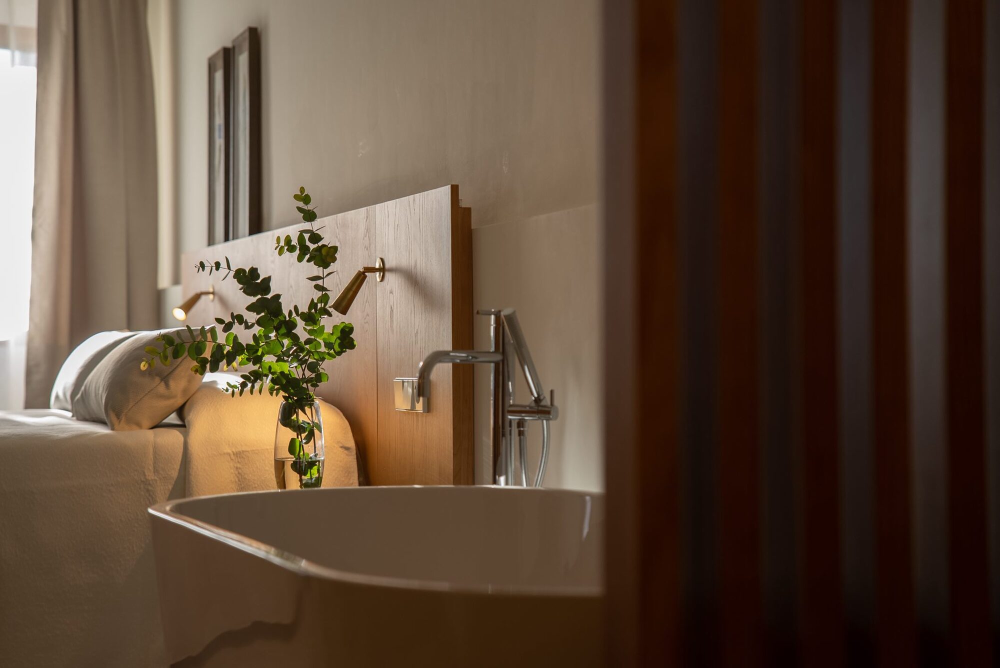 A NEMA room detail: an oak headboard with a brass reading light, a glass vase of fresh eucalyptus, white linen and a freestanding bath just in view