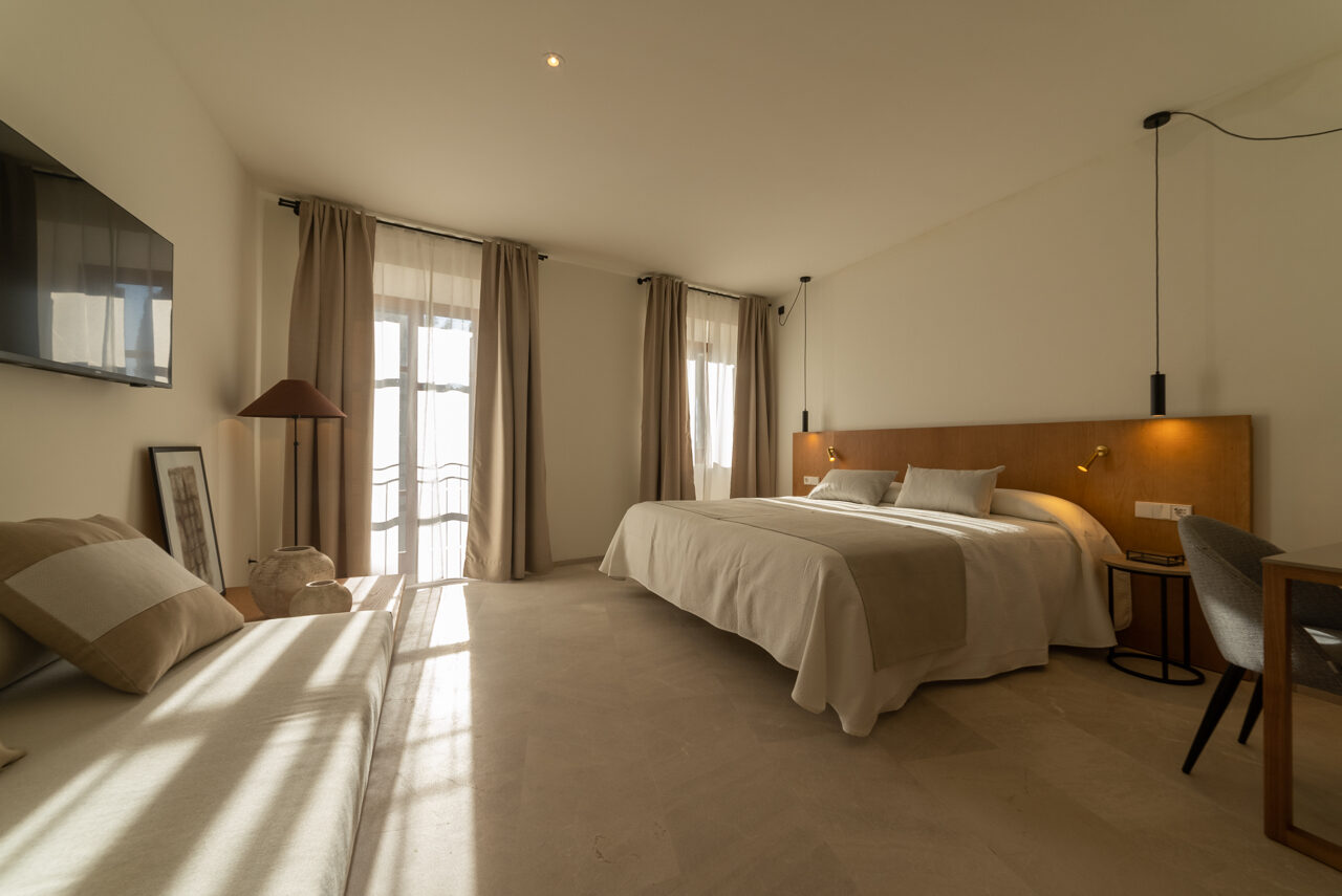 A NEMA double room with high ceilings, an oak headboard, warm pendant and bedside lights, a clay lamp and curtained French windows letting a soft band of light across the stone floor