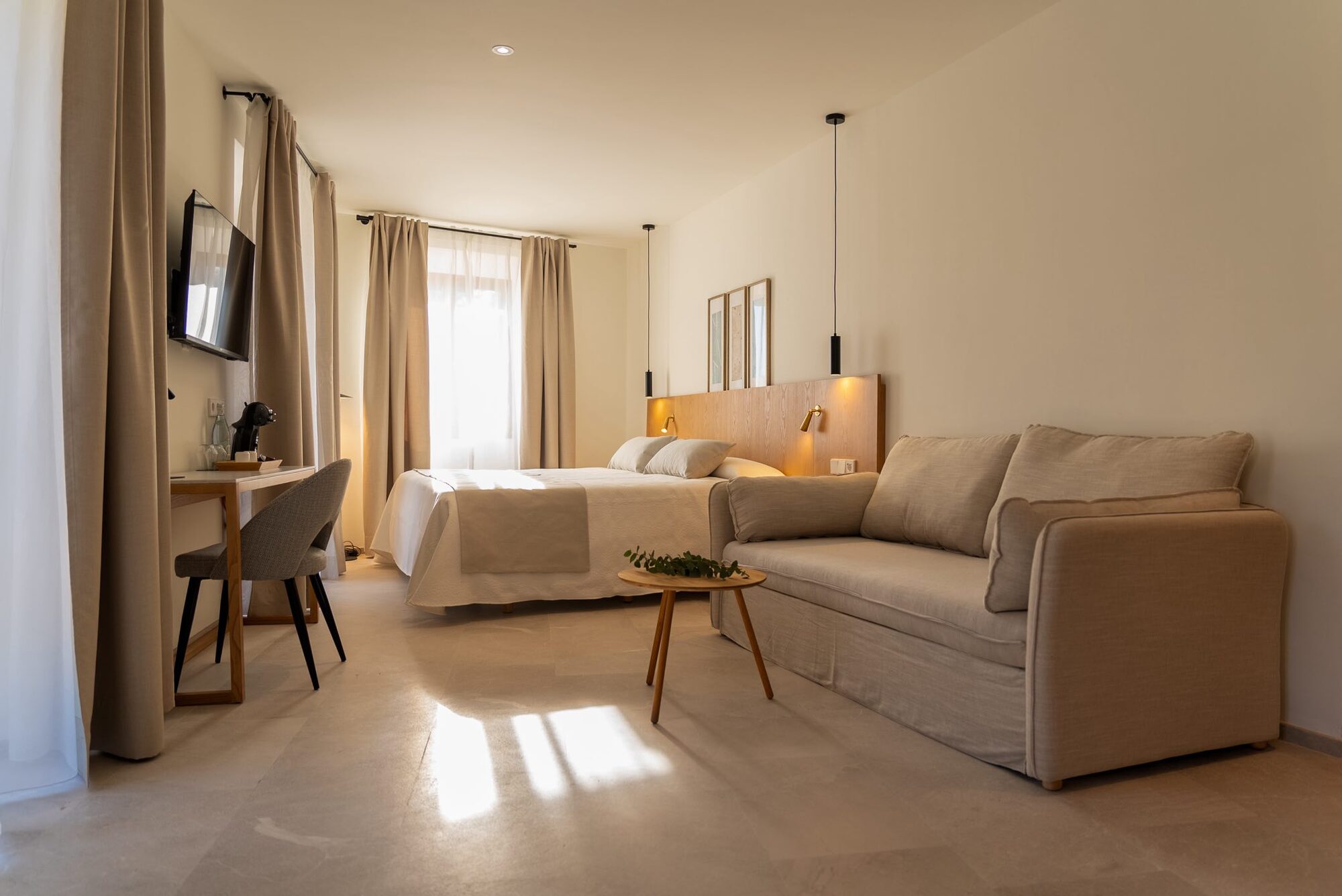 A bright NEMA suite with a pale oak headboard and reading lights, a long linen sofa, a small round oak table with eucalyptus, and tall windows onto the street
