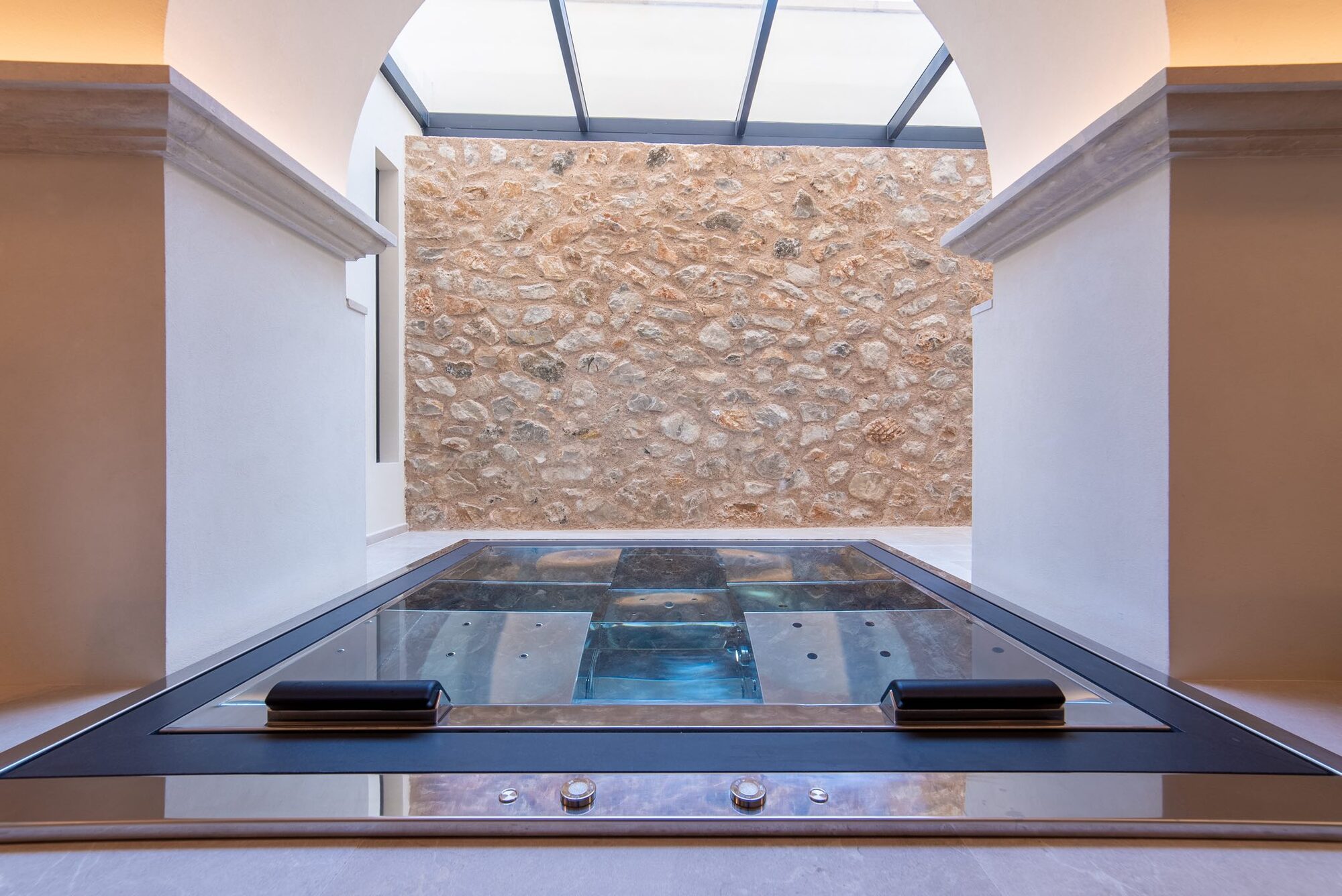 The NEMA spa: a steel hydrotherapy pool set into pale stone floor beneath a glazed roof, facing a tall wall of rough golden marès stone in two arched alcoves