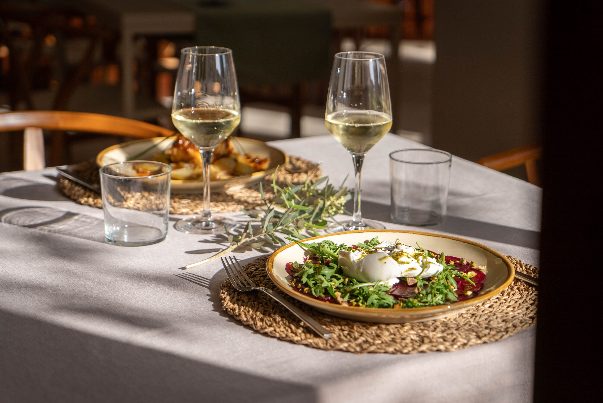 A NEMA table laid in low evening light: linen cloth, woven placemats, two glasses of white wine, a sprig of olive across the cloth and a plate of beetroot and burrata
