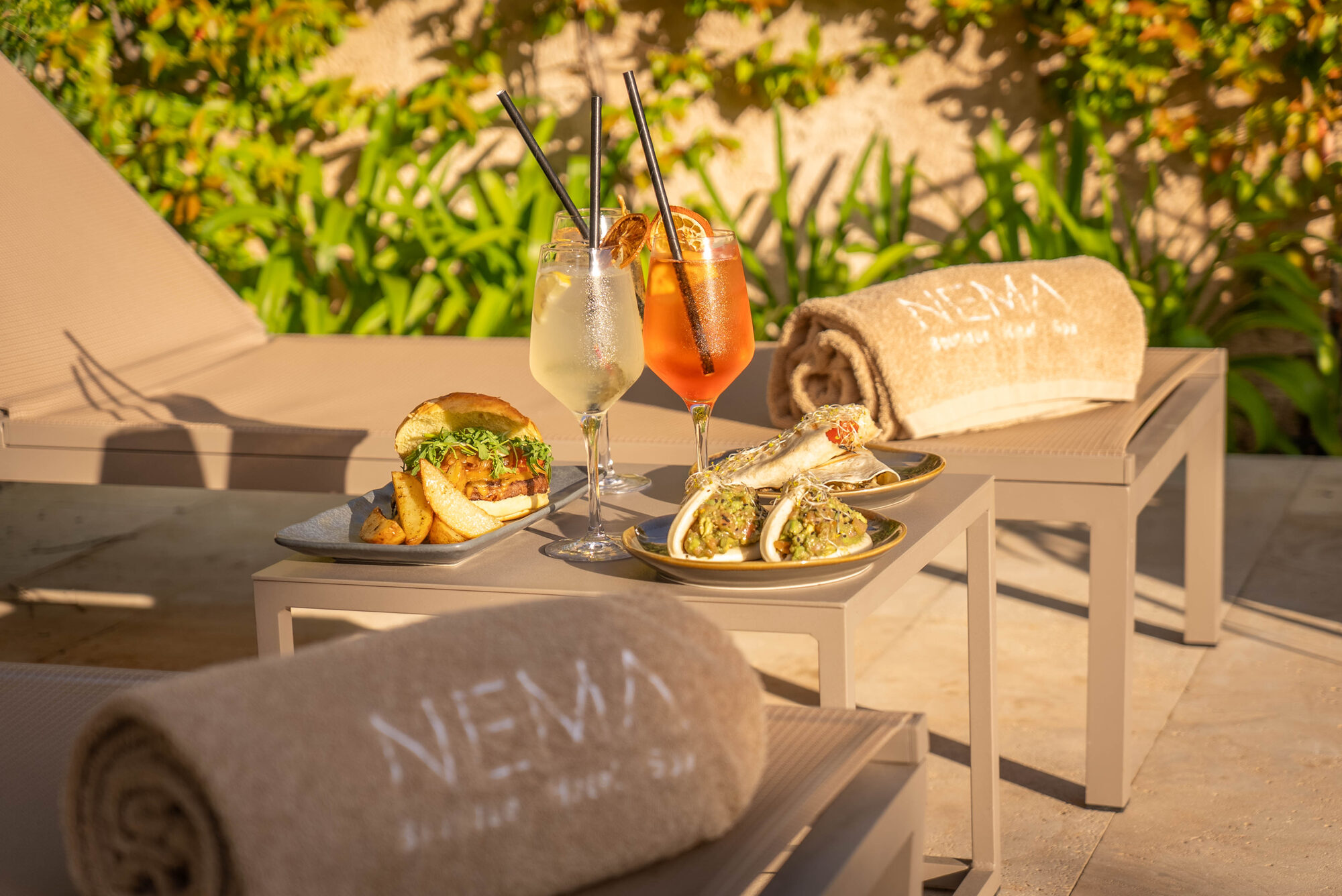 Two loungers in the NEMA garden set with rolled NEMA-embroidered towels, two spritz cocktails and small plates against a bank of orange flowers